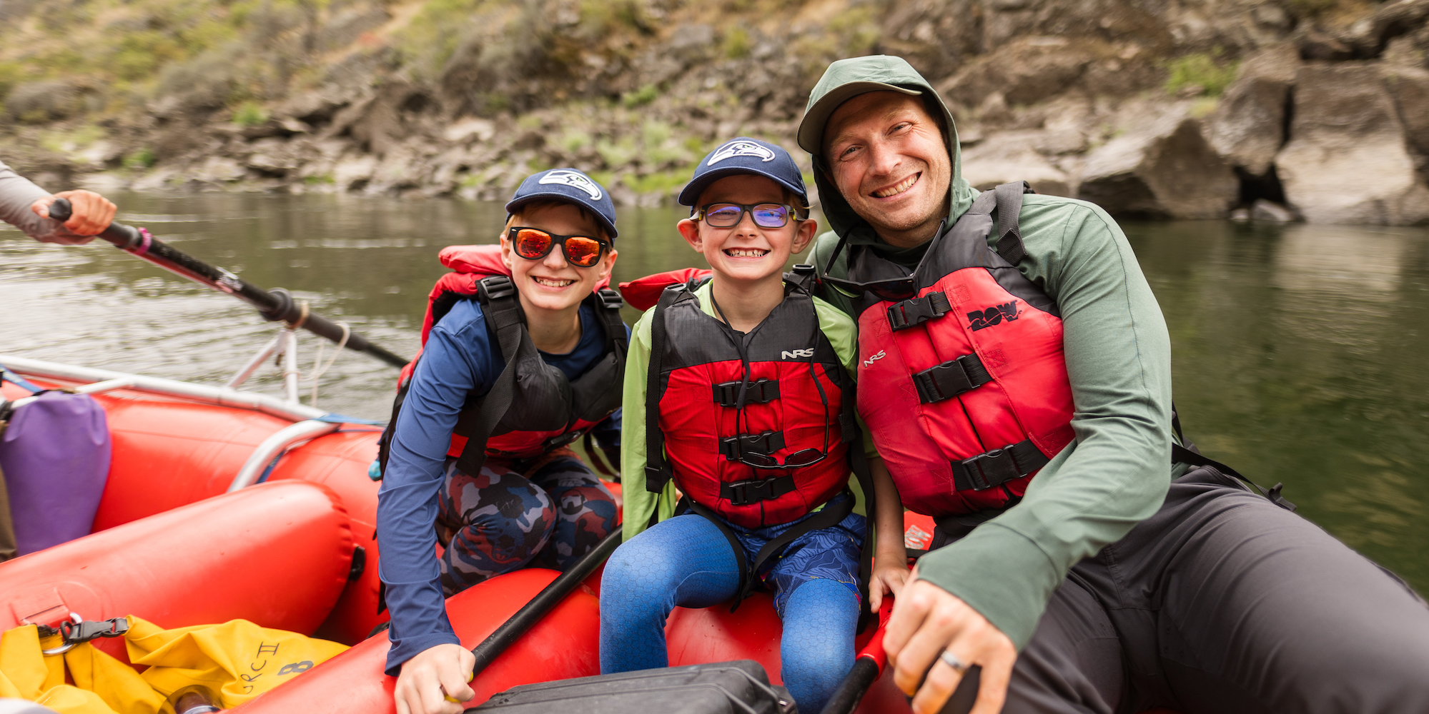 A dad and his two sons sitting on a red whitewater raft smiling together