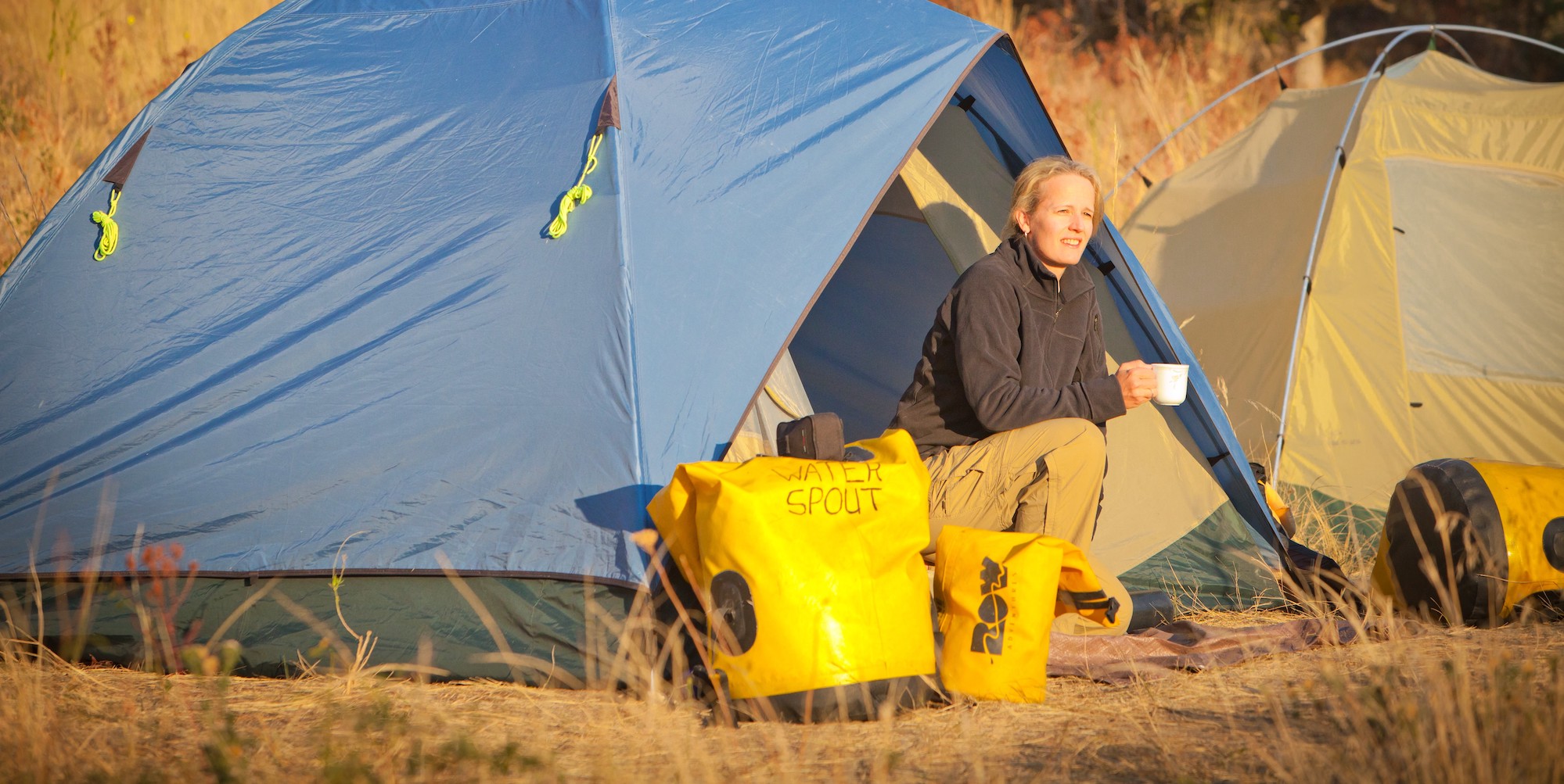 A woman sitting outsider her blue tent at sunset with numerous yellow dry bags outside her tent