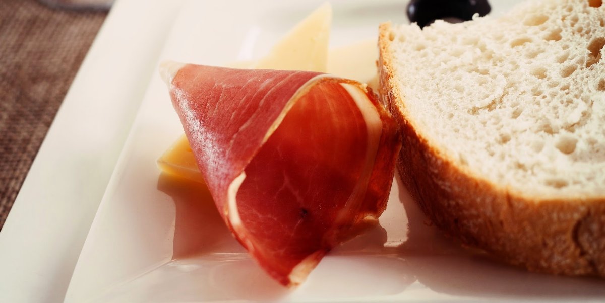 Prosciutto on a white plate next to a slice of bread 