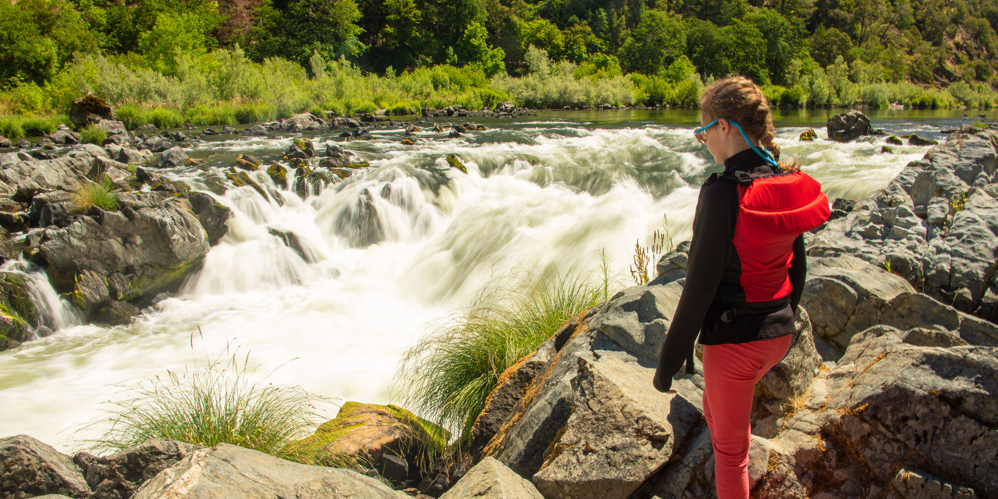 A girl standing along the Rogue River over looking a rapid with her red PFD on and chums with sunglasses around her head.