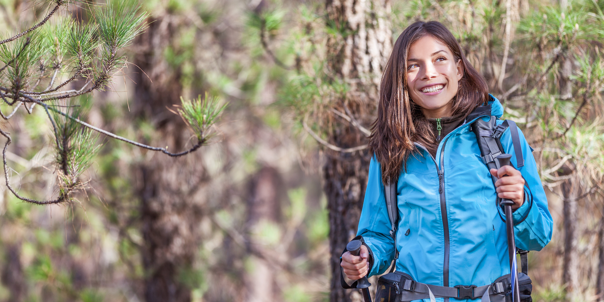 A girl in a blue rain jacket holding hiking poles smiling up at the towering pine trees that surround her while on a hike