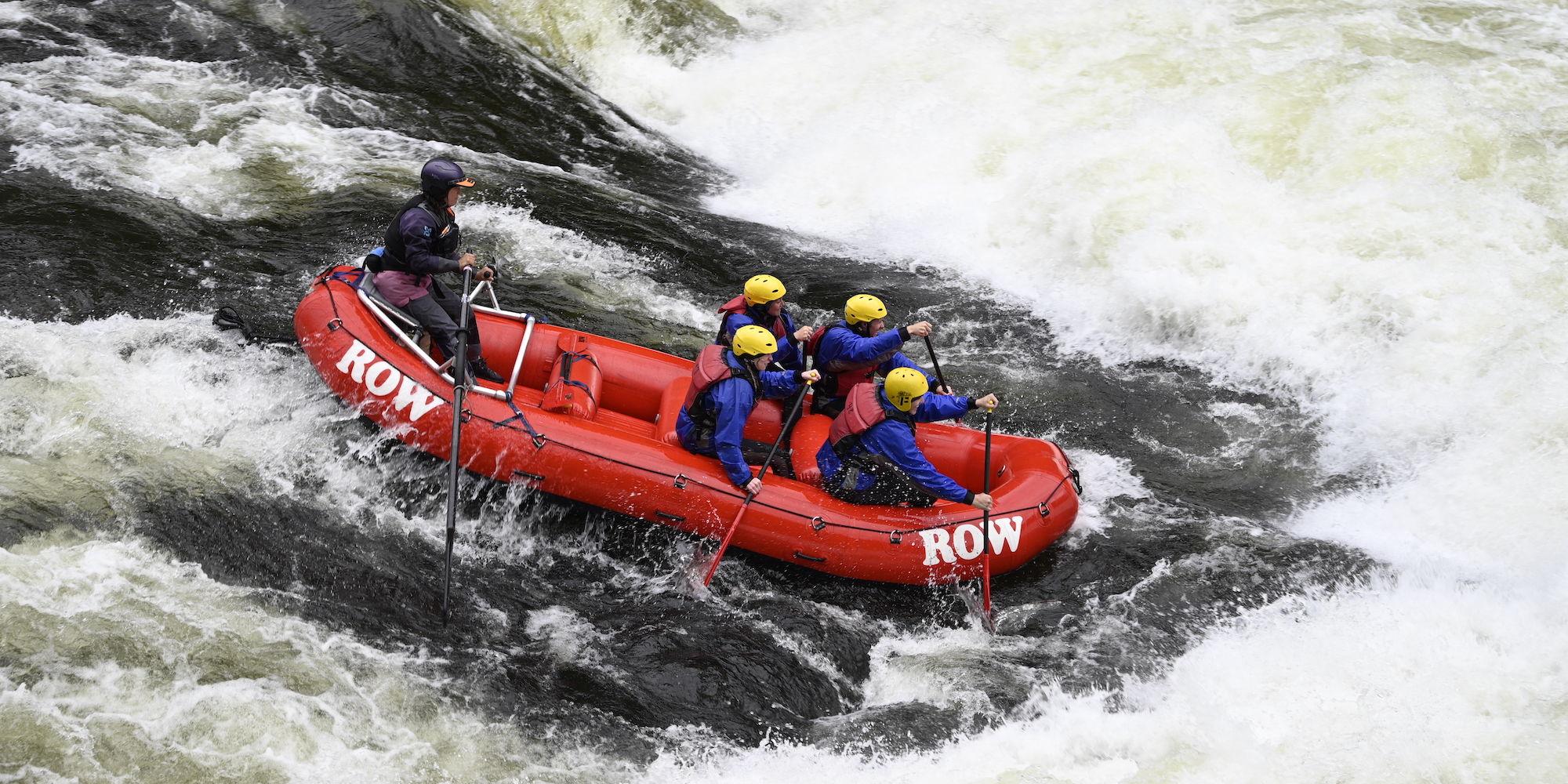 A red raft full of paddlers and a guide on a stern frame in the back paddling through a rapid on the Lochsa River