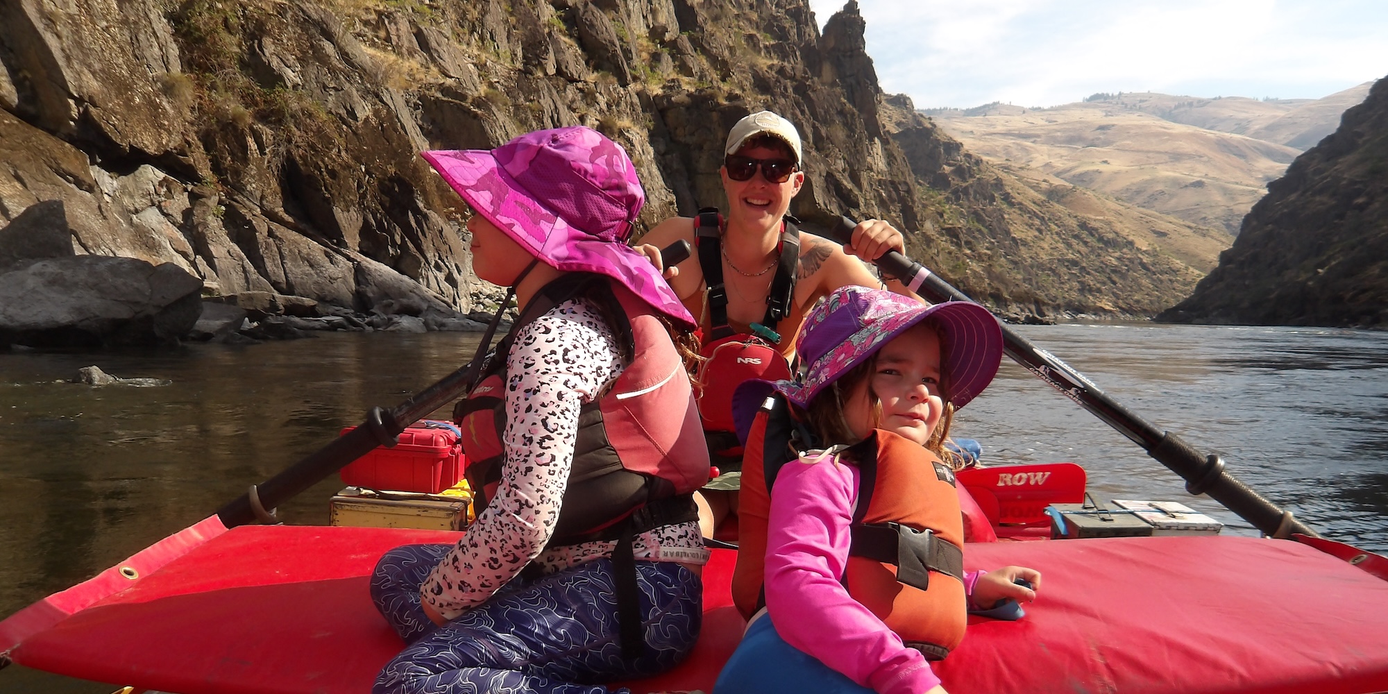 Two kids sitting on a red ROW Adventures raft while their guide rows them downstream.