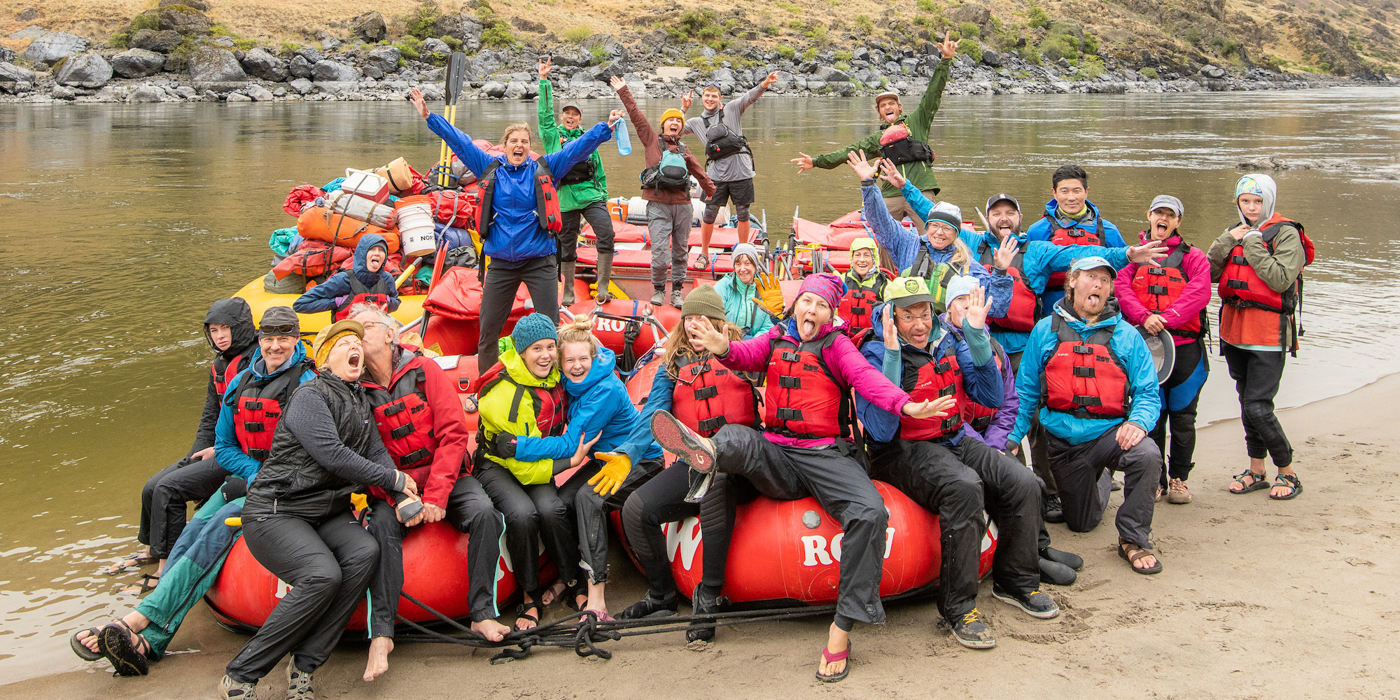 A group of people sitting across a couple red whitewater rafting smiling silly on the river