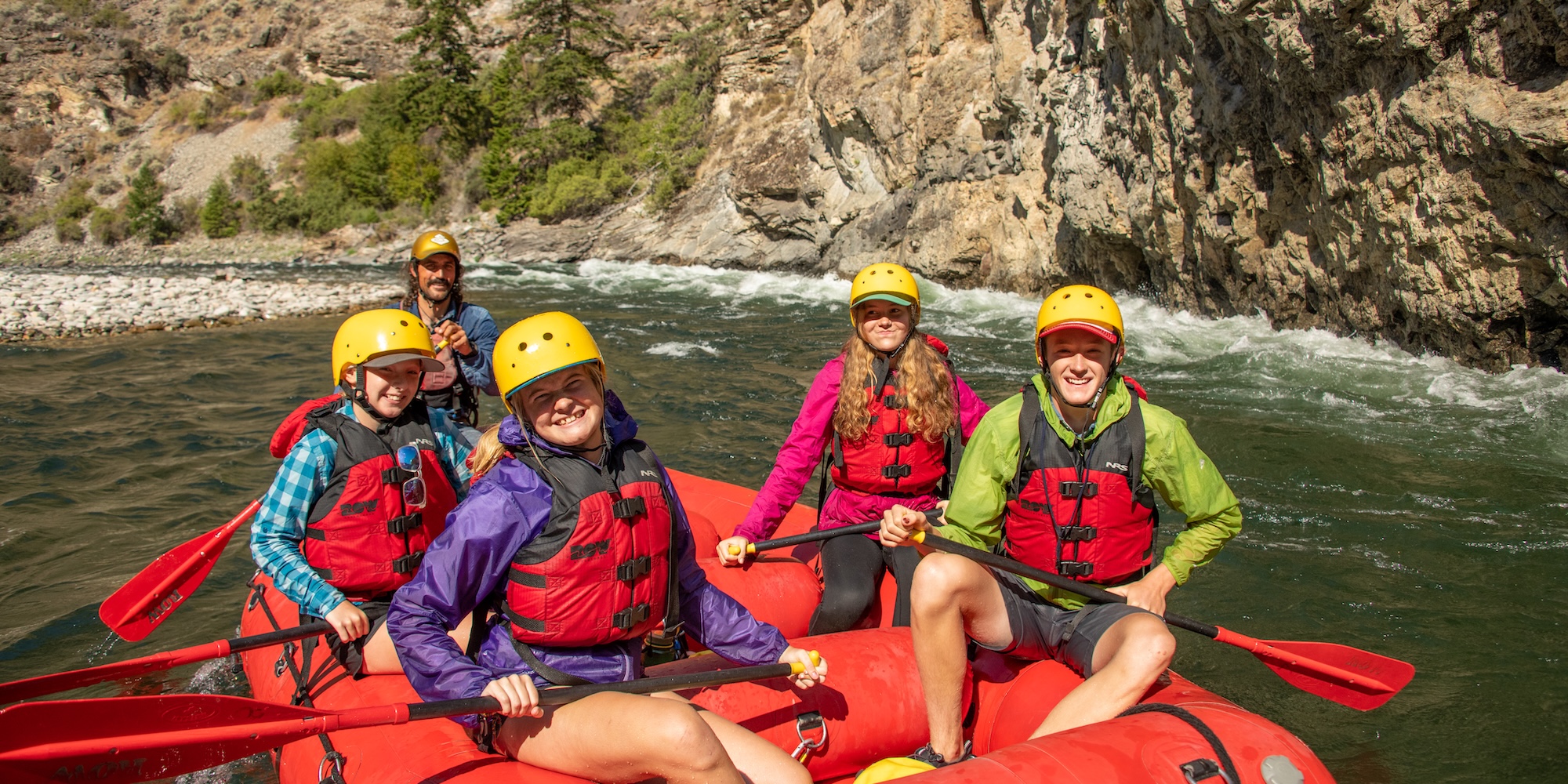 Group of smiling rafters in yellow helmets and red life jackets floating past a cliffside on the Middle Fork of the Salmon River.