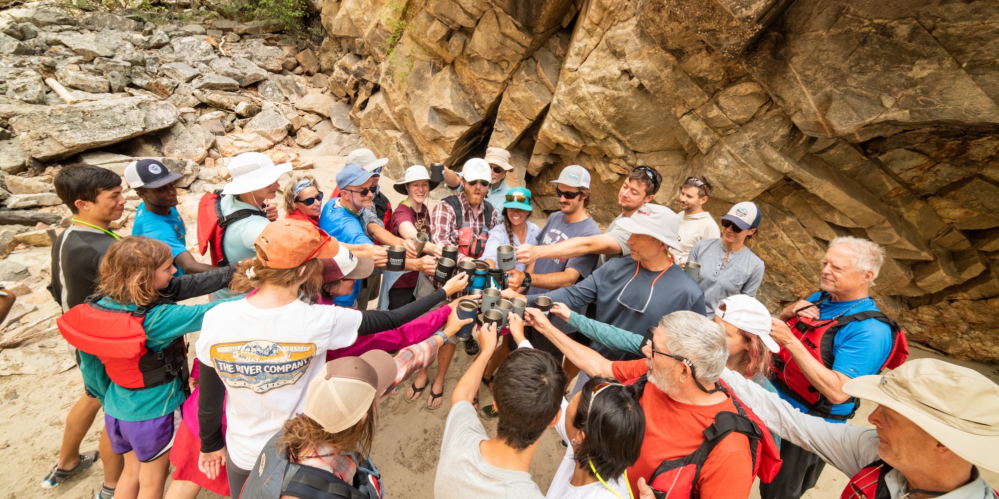 Group of rafters gathered on the riverbank raising mugs in a celebratory toast, surrounded by canyon walls during a guided Salmon River trip.
