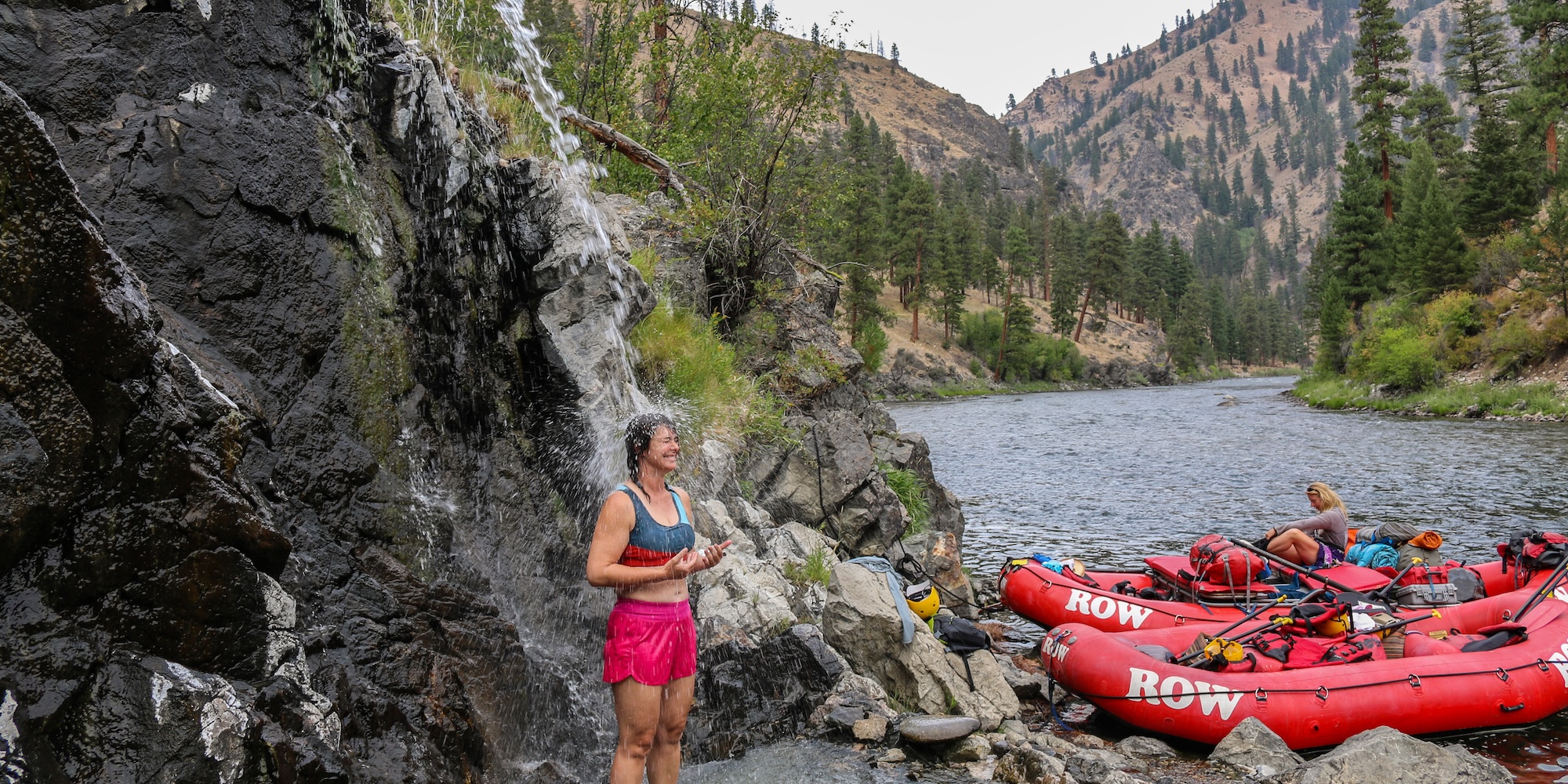 Rafter standing beneath a natural waterfall shower beside the Middle Fork of the Salmon River, with rafts pulled ashore during a quiet moment on the trip.