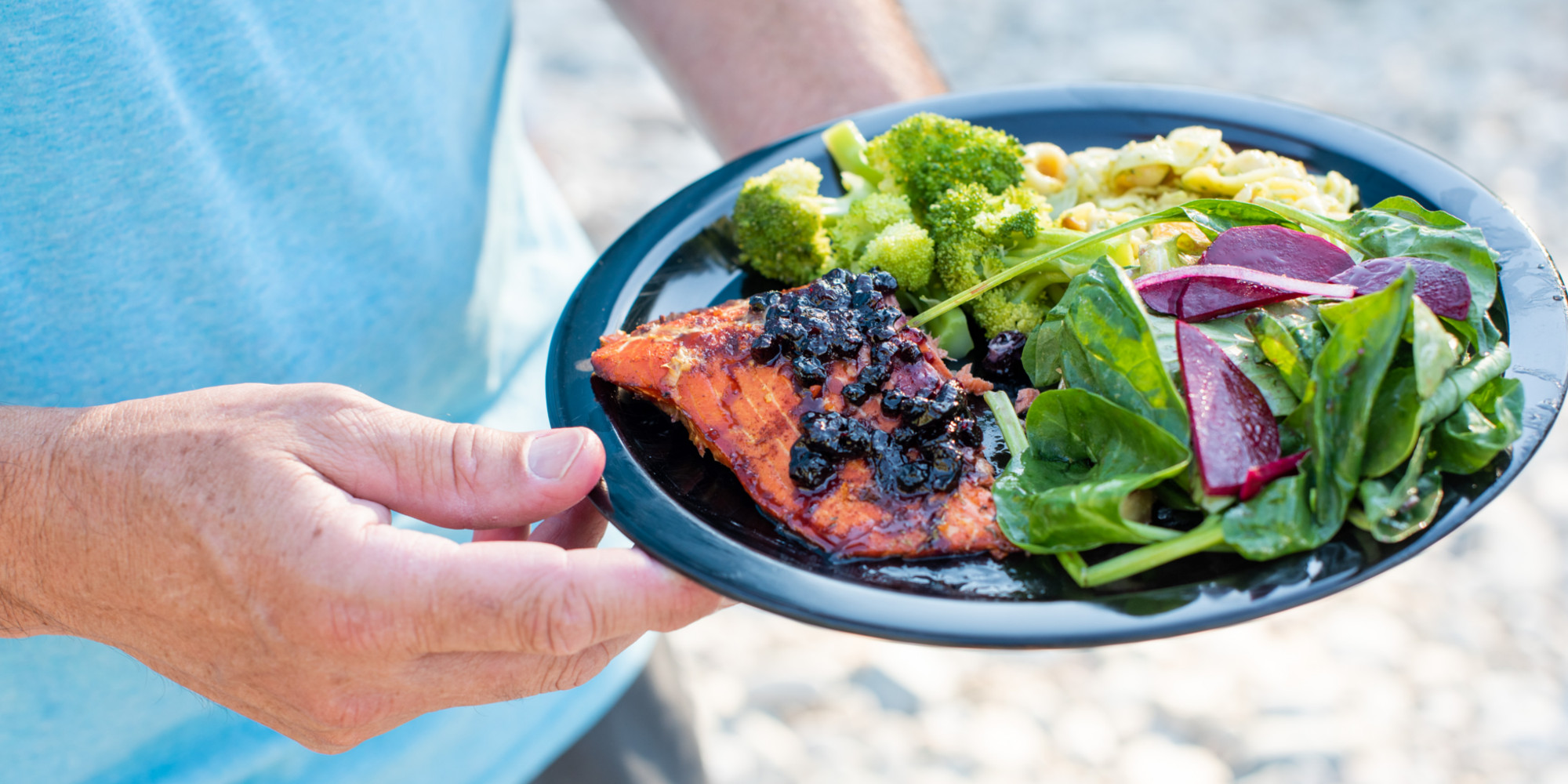A hand holding a black plate of cooked salmon with huckleberries on top and a salad on the side