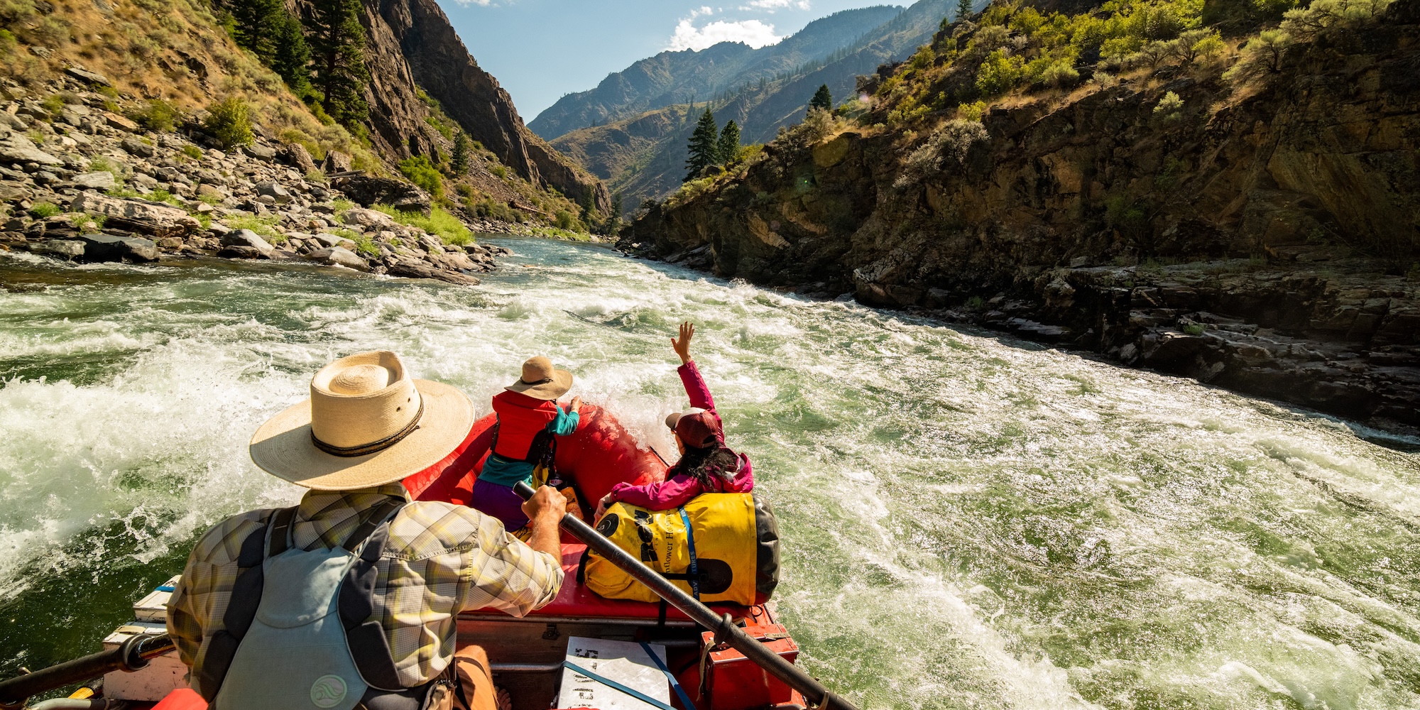 Guests in a red raft riding through a scenic canyon on the Middle Fork of the Salmon River, raising their hands as they navigate whitewater surrounded by dramatic mountain views.