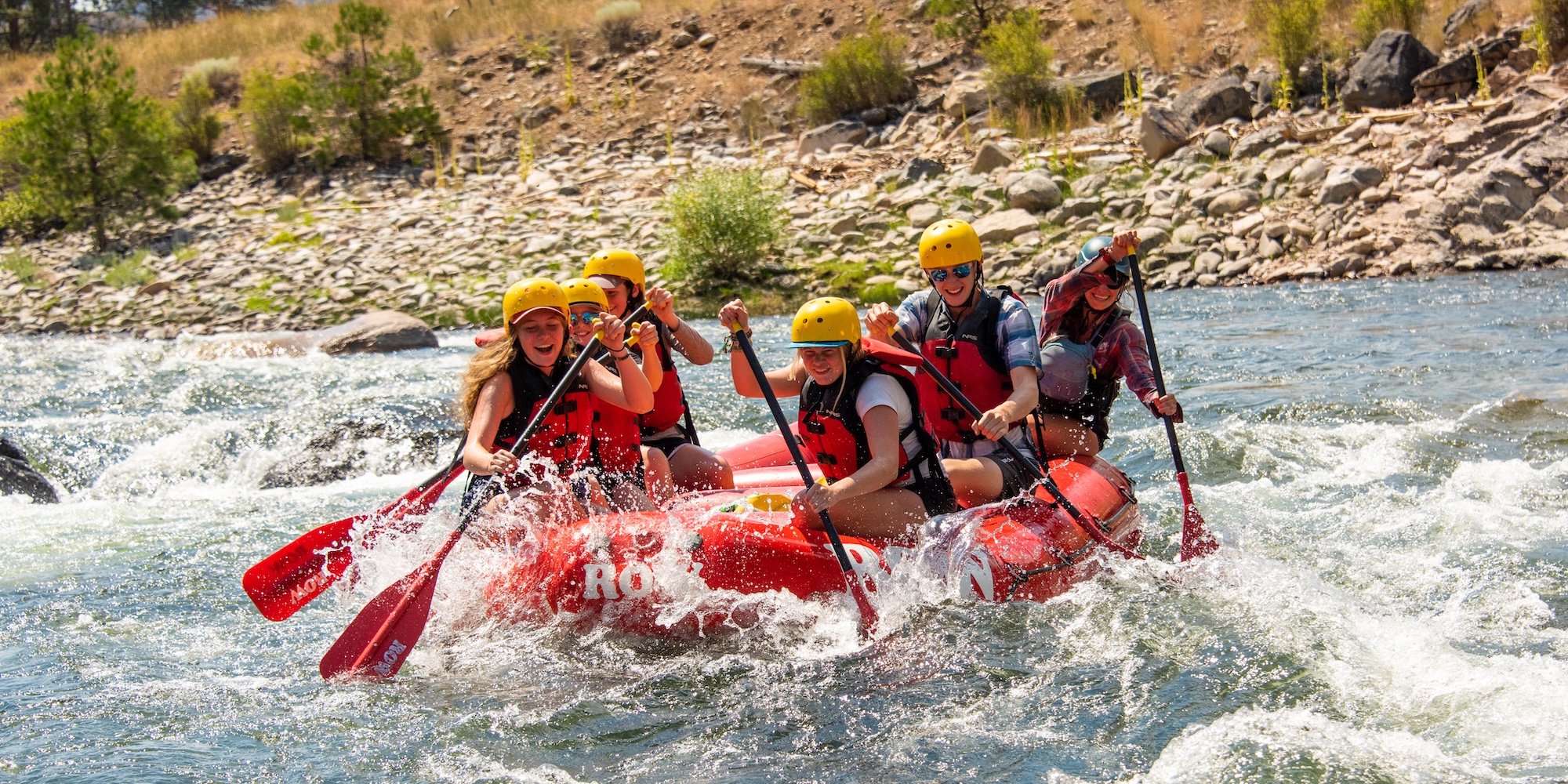 Smiling group of paddlers in a red raft charging through whitewater on the Middle Fork of the Salmon River, wearing helmets and life jackets.