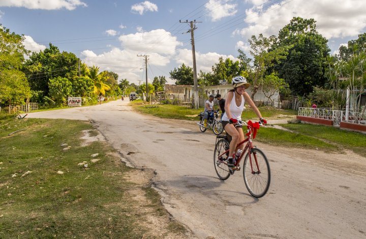 A woman on a bike riding on a paved road in Cuba with green grass and trees behind her