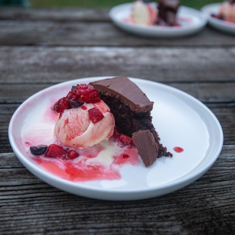 Raspberry chocolate cake with a scoop of vanilla ice cream on a white plate on an outdoor picnic table