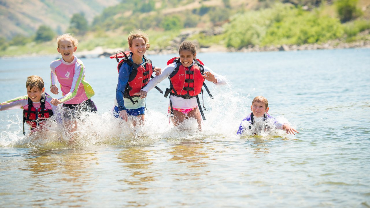 Group of kids all in life jackets running through the Salmon River and smiling on a sunny summer day