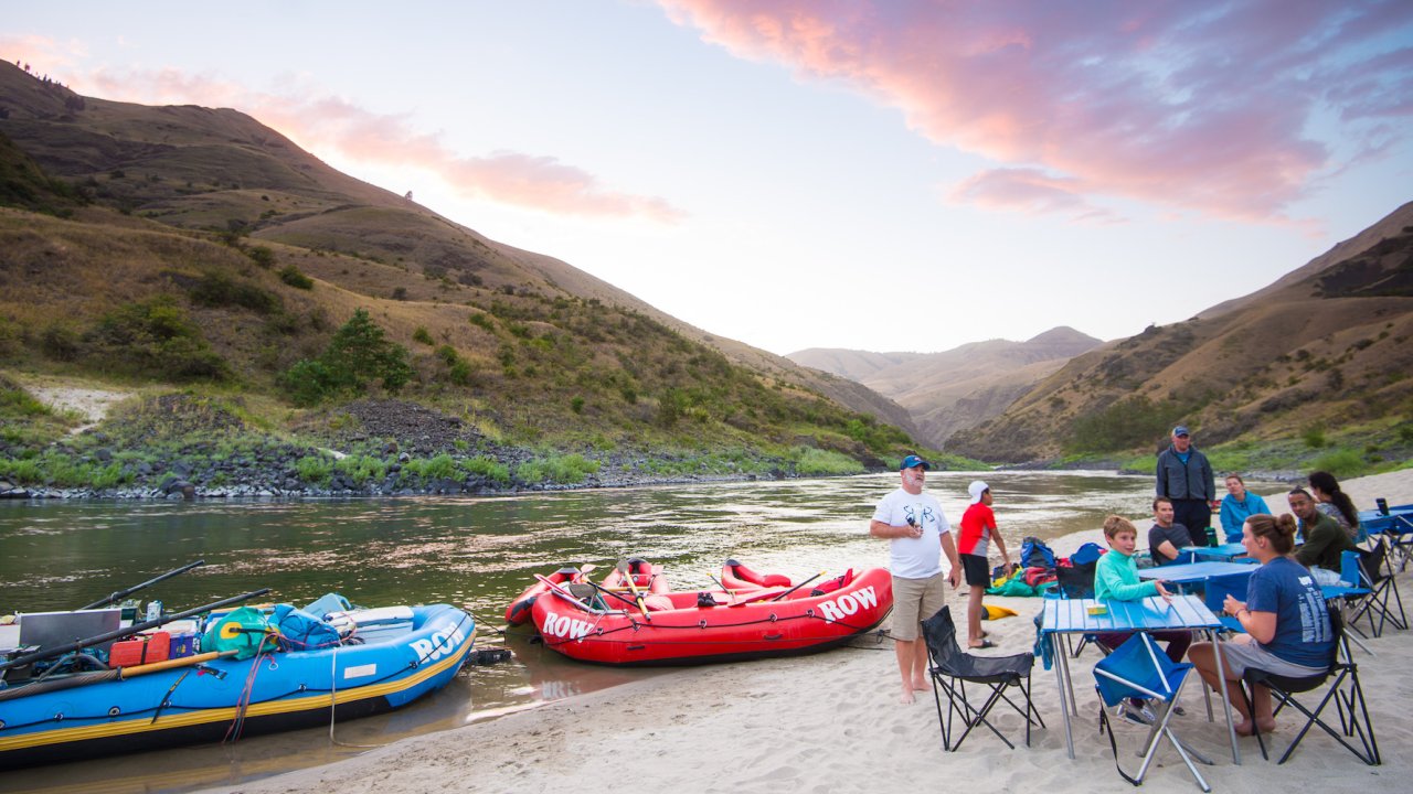 Family sitting around blue tables on a beach next to whitewater rafts as the sunsets hue purple and pink in the sky above