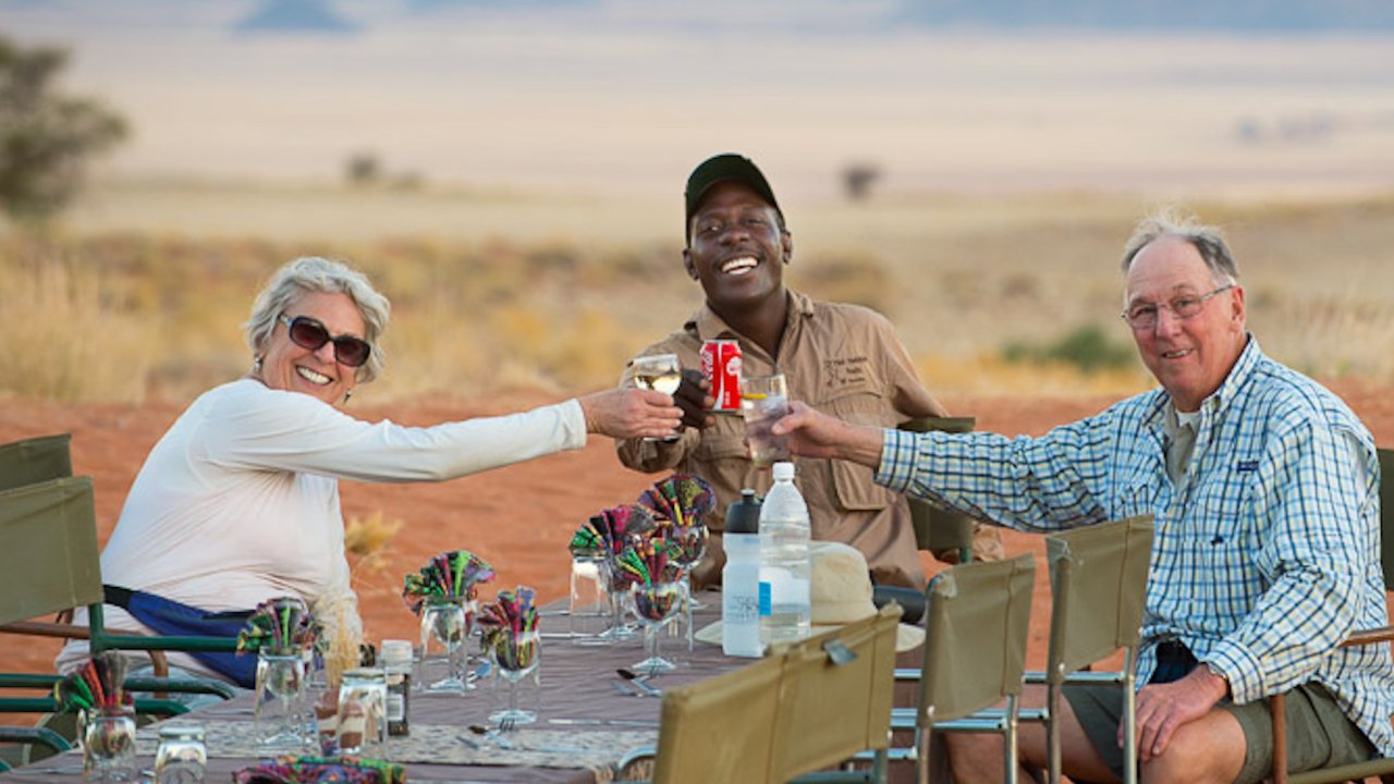 Two tourists and an African safari guide sit around a table and cheers their soda to a great day