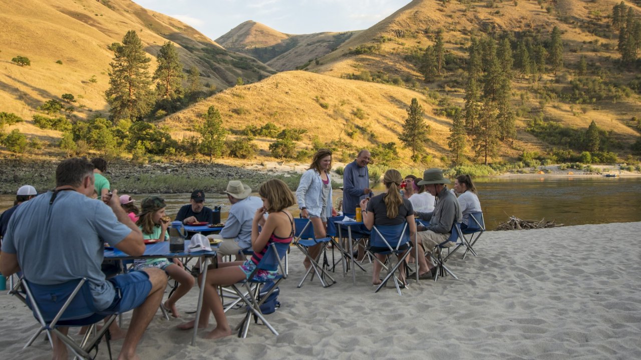 Group of people enjoying a meal on a sandy beach next to a river.