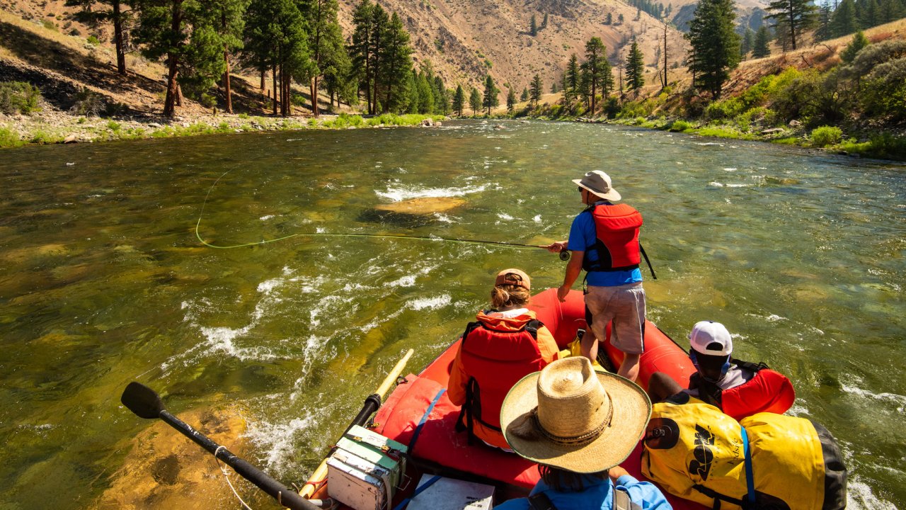 Person standing up in a red raft to cast a fly fishing line while a guide rows them down the river in Idaho