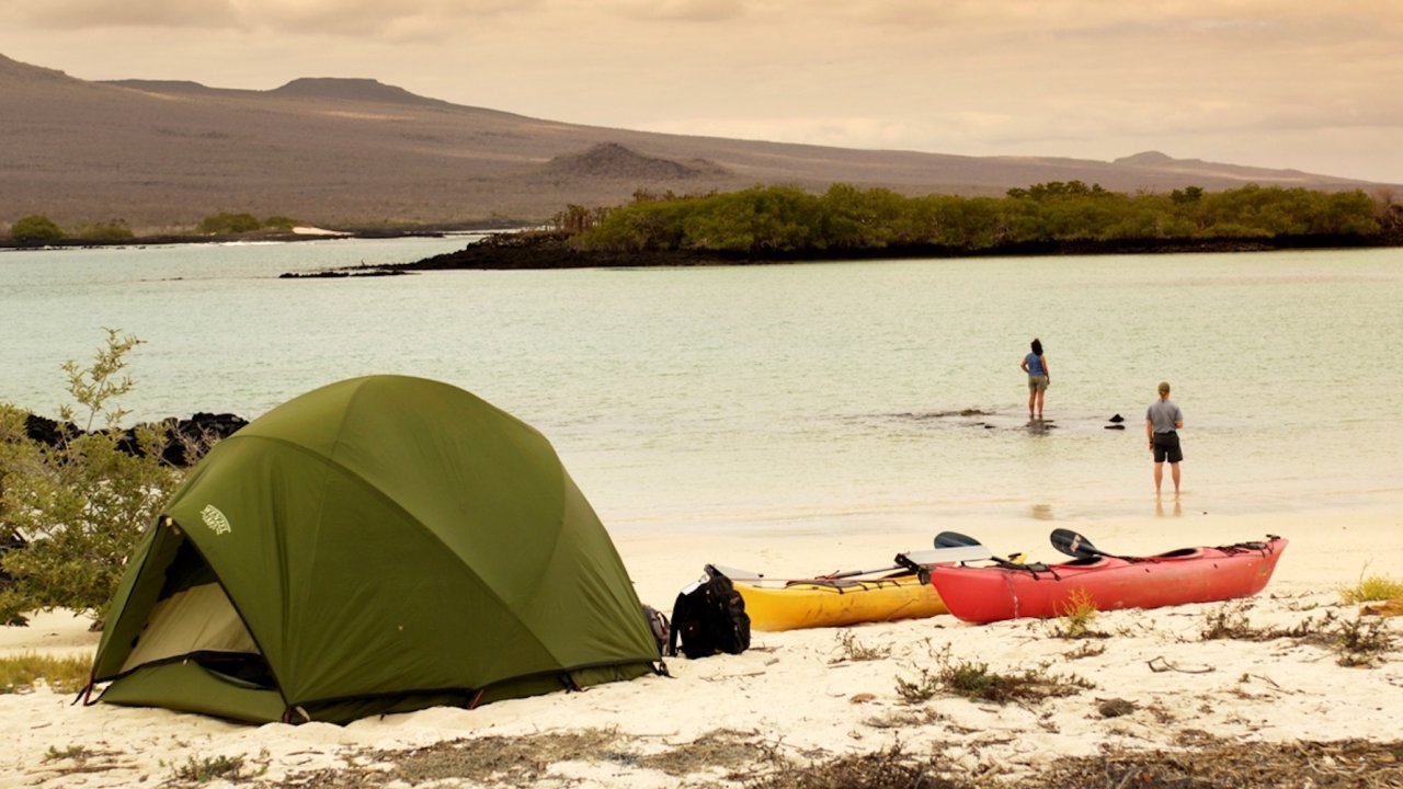 A couple dipping their feet in the water on a beach in the Galapagos with their tent behind them