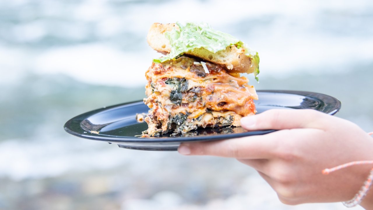 Hand holding a plate of lasagna and garlic bread by the river.