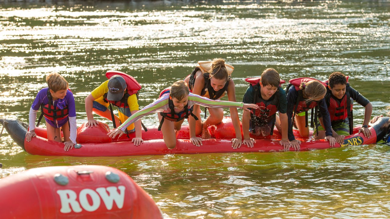 A row of kids on top of an upside down inflatable kayak in the Salmon River