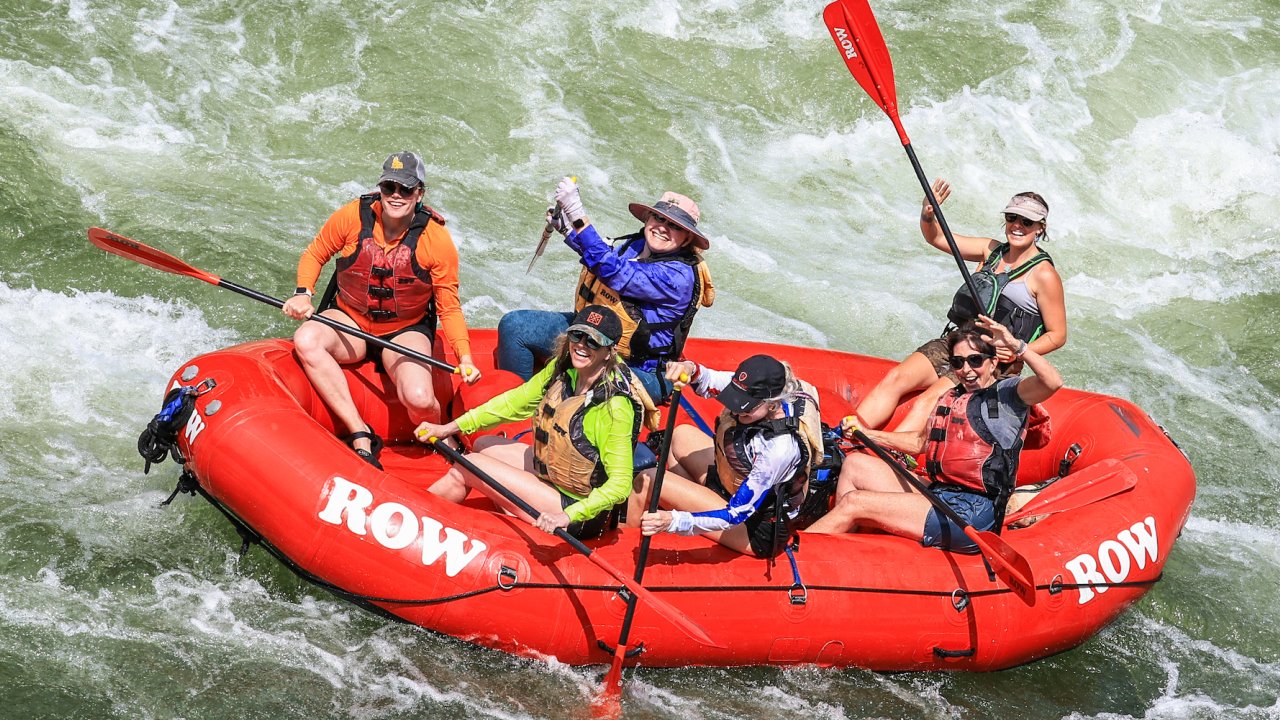 A red ROW branded raft full of people holding red and black paddles with the guide in the back holding their paddle up in the air