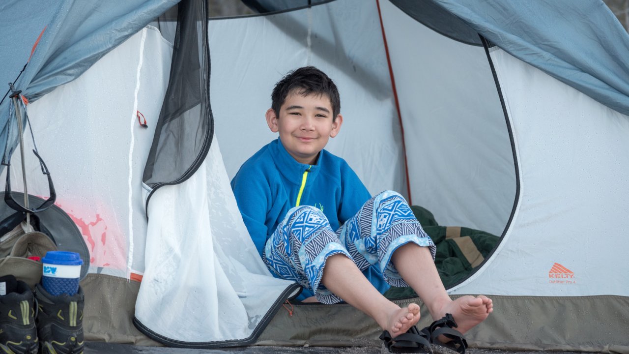 A boy sitting out the zippered door to his tent while camping