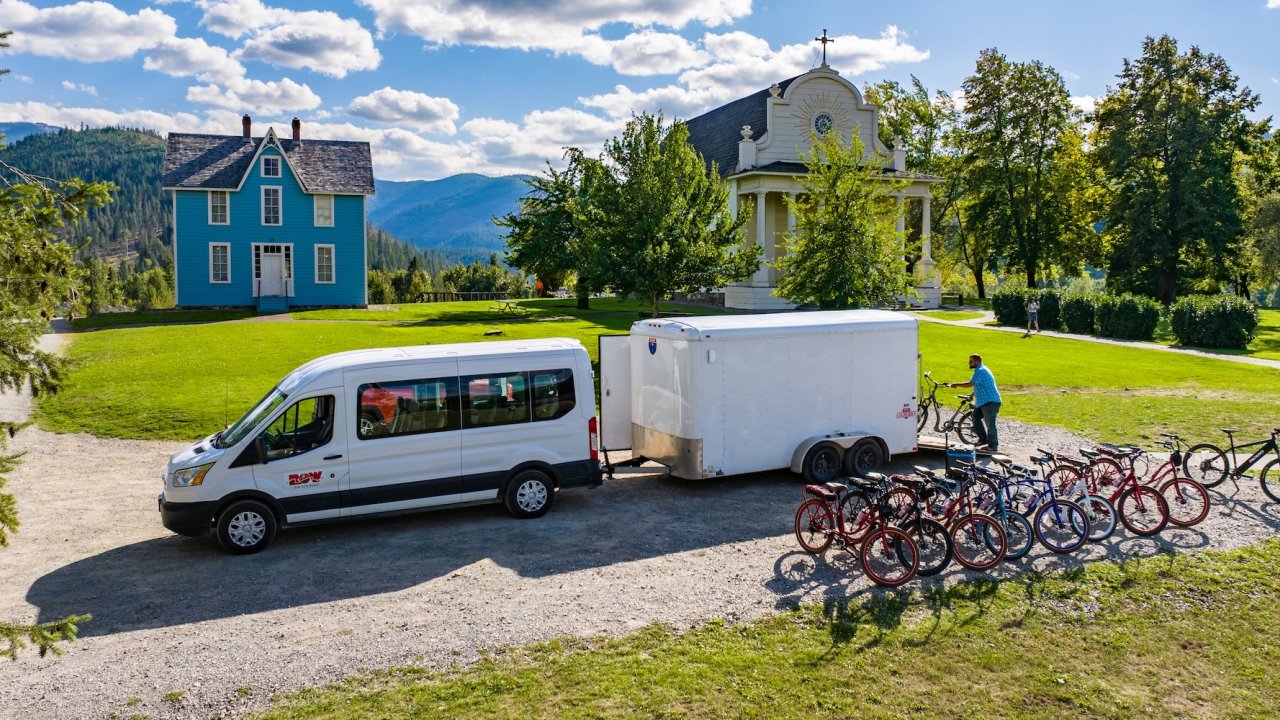 ROW Adventures support van and bike trailer parked near historic buildings in a scenic Idaho mountain setting, with guides unloading bikes for a cycling tour.