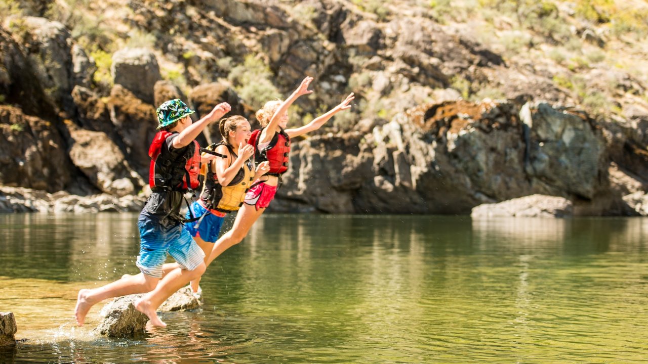Kids in life jackets jumping together into the river from a shallow rocky edge.