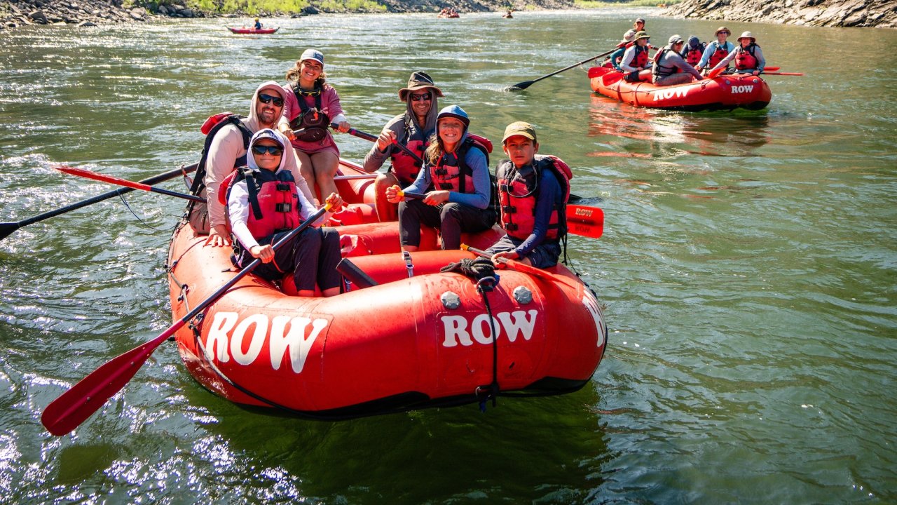 Smiling guests in red life jackets paddling an oar raft through a sunny canyon stretch while a ROW raft floats behind them.