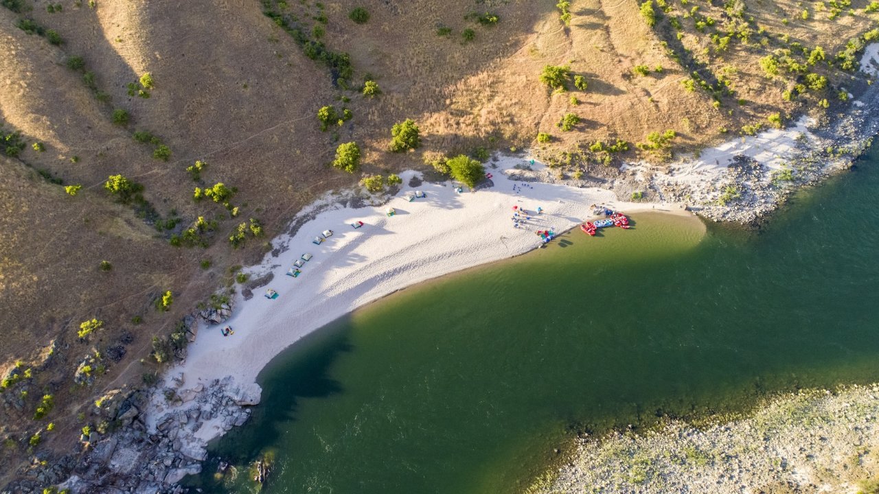 Aerial view of sandy riverside camp with tents and rafts along a clear green river.