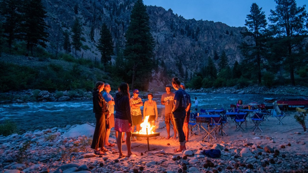 Guests gathered around a campfire on a sandy riverside at dusk.