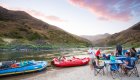 Family sitting around blue tables on a beach next to whitewater rafts as the sunsets hue purple and pink in the sky above