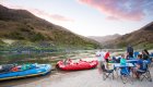 Beached whitewater rafts next to people sitting at a camping table enjoying the sunset along the river