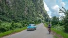 A person biking on the side of a road away from the camera as an old blue car passes by in Cuba