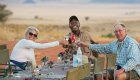 Two tourists and an African safari guide sit around a table and cheers their soda to a great day 