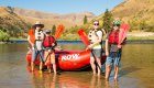 A family standing in a river holding rafting paddles while wearing life jackets smiling in front of their whitewater raft