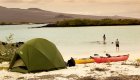 A couple dipping their feet in the water on a beach in the Galapagos with their tent behind them