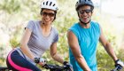 Two people on bikes smiling with helmets and sunglasses on with green trees behind them