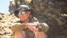 Man sitting along a river bank wearing sunglasses and a hat