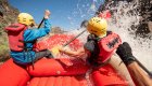The back of two people wearing ROW Adventures life jackets as they get splashed while paddling on a red raft through a whitewater rapid