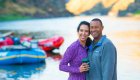 A couple smiling and holding drinks while standing in the sand in front of whitewater rafts floating in the Salmon River in Idaho