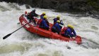 Whitewater rafting guide on a stern frame rowing through a whitewater rapid with passengers paddling 