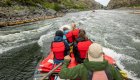 Picture taken from the back of a red whitewater raft looking downstream as someone rows with passengers in the front