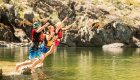 Kids in life jackets jumping together into the river from a shallow rocky edge.