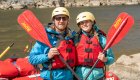 Two guests smiling while holding red ROW branded paddles and wearing yellow helmets about to start whitewater rafting