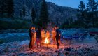 Guests gathered around a campfire on a sandy riverside at dusk.