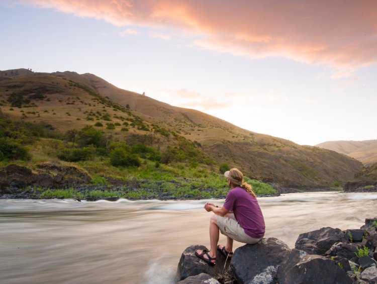 A ROW guest sitting peacefully on river rocks, watching the flowing water at sunset.
