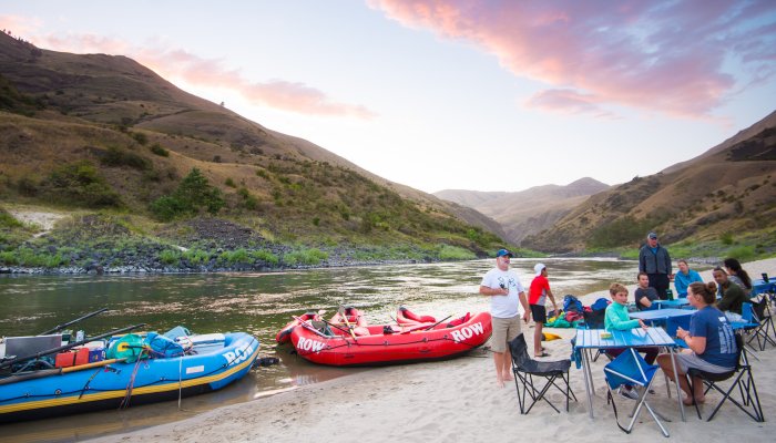 A group of people camping sitting around a blue camp table on a sandy beach next to red and blue rafts on the Lower Salmon River in Idaho