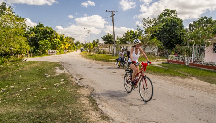 A woman on a bike riding on a paved road in Cuba with green grass and trees behind her