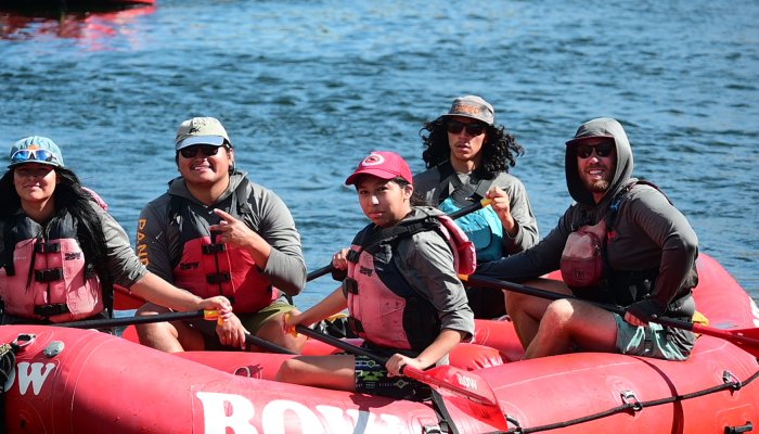 Young participants from the Pandion Institute rafting program paddle a red ROW raft across calm river water, learning river skills and teamwork under blue skies.